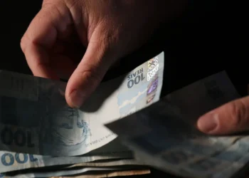 Mãos contam cédulas de cem reais sobre fundo escuro em close que evidencia o dinheiro em espécie