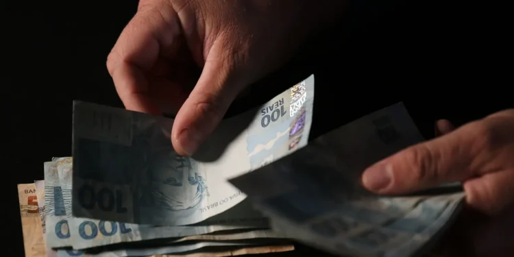 Mãos contam cédulas de cem reais sobre fundo escuro em close que evidencia o dinheiro em espécie