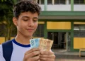 Estudante segura notas de dinheiro em frente à escola, representando os pagamentos de até R$ 1.000 do Pé-de-Meia em maio de 2026.