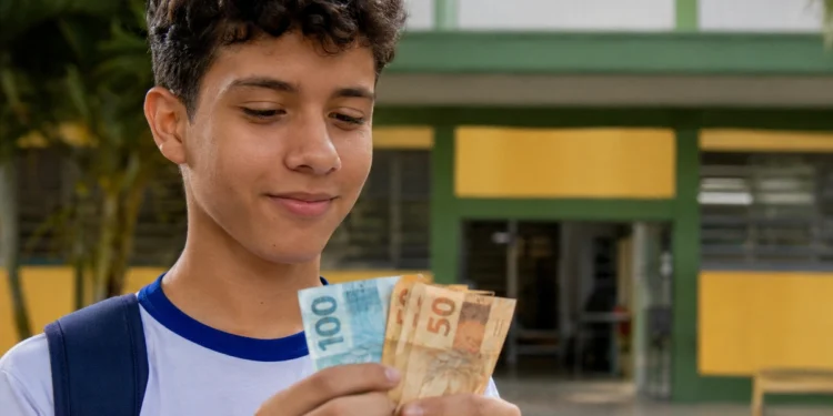 Estudante segura notas de dinheiro em frente à escola, representando os pagamentos de até R$ 1.000 do Pé-de-Meia em maio de 2026.