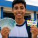 Jovem sorridente segura notas de R$ 100 e cartão do programa Pé-de-Meia em frente a escola pública.