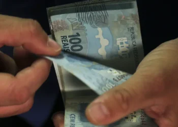 Mãos contam cédulas de cem reais sobre fundo escuro.