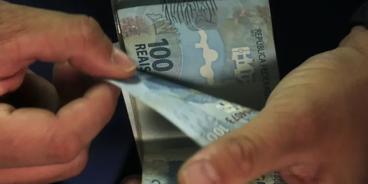Mãos contam cédulas de cem reais sobre fundo escuro.