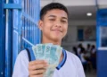 Estudante de escola pública segurando notas de R$ 100 em ambiente escolar representa o pagamento do Pé-de-Meia de R$ 200 e R$ 225 em maio.