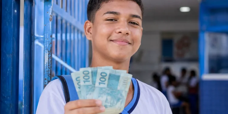 Estudante de escola pública segurando notas de R$ 100 em ambiente escolar representa o pagamento do Pé-de-Meia de R$ 200 e R$ 225 em maio.
