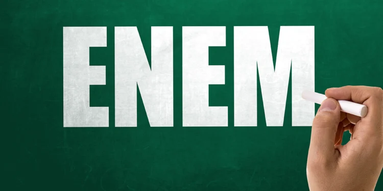 Mão com giz em frente a quadro verde com a palavra Enem em destaque, representando o período de recursos para isenção do Enem 2026.