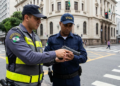 Agente de trânsito com colete amarelo e vigilante com boné azul examinam pistola em cruzamento urbano.