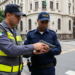 Agente de trânsito com colete amarelo e vigilante com boné azul examinam pistola em cruzamento urbano.