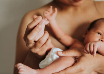 Mãe segura o bebê recém-nascido nos braços, com a mãozinha da criança apoiada em sua mão.