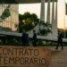 Placa de madeira com a inscrição contrato temporário em primeiro plano diante de prédio público e bandeiras do Brasil ao fundo