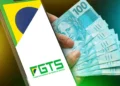 Smartphone exibindo logotipo do FGTS ao lado de cédulas de cem reais sobre fundo verde e amarelo com bandeira do Brasil