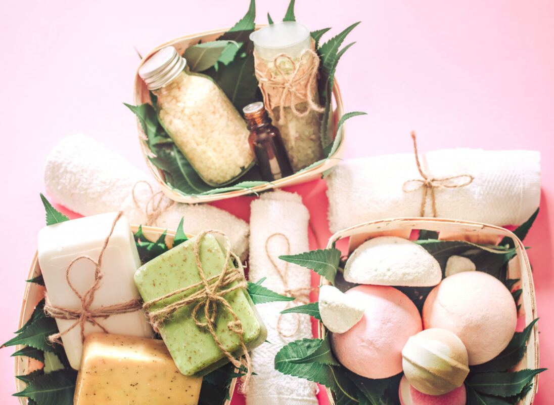 Cestas com sabonetes artesanais, bombas de banho, sais e toalhas sobre fundo rosa