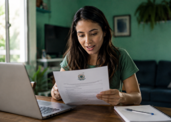 Jovem mulher de cabelos castanhos lendo documento oficial com brasão brasileiro, sentada em mesa com notebook cinza.