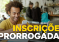 Jovem negro sorridente de cabelo crespo e camiseta amarela olha celular em cafeteria com notebook aberto