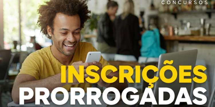 Jovem negro sorridente de cabelo crespo e camiseta amarela olha celular em cafeteria com notebook aberto