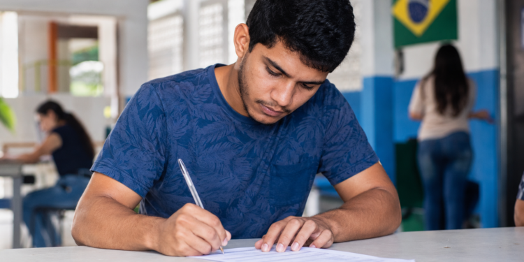 Jovem de camiseta azul escrevendo em folha de prova com bandeira do Brasil ao fundo em ambiente escolar.