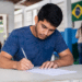 Jovem de camiseta azul escrevendo em folha de prova com bandeira do Brasil ao fundo em ambiente escolar.