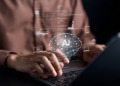 Mãos digitando em laptop com holograma de cérebro AI e ícones de tecnologia flutuando sobre teclado.