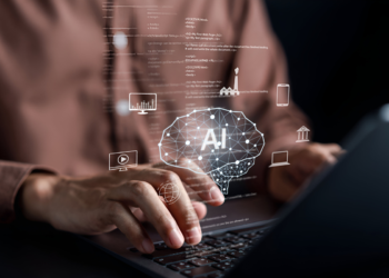 Mãos digitando em laptop com holograma de cérebro AI e ícones de tecnologia flutuando sobre teclado.