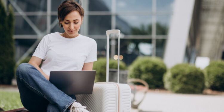 Profissional trabalhando no notebook durante viagem com mala ao lado em área externa de aeroporto.