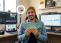 Homem sorrindo segura maço de notas de 100 reais em estúdio com monitores, ring lights, câmeras e placas do YouTube