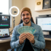 Homem sorrindo segura maço de notas de 100 reais em estúdio com monitores, ring lights, câmeras e placas do YouTube