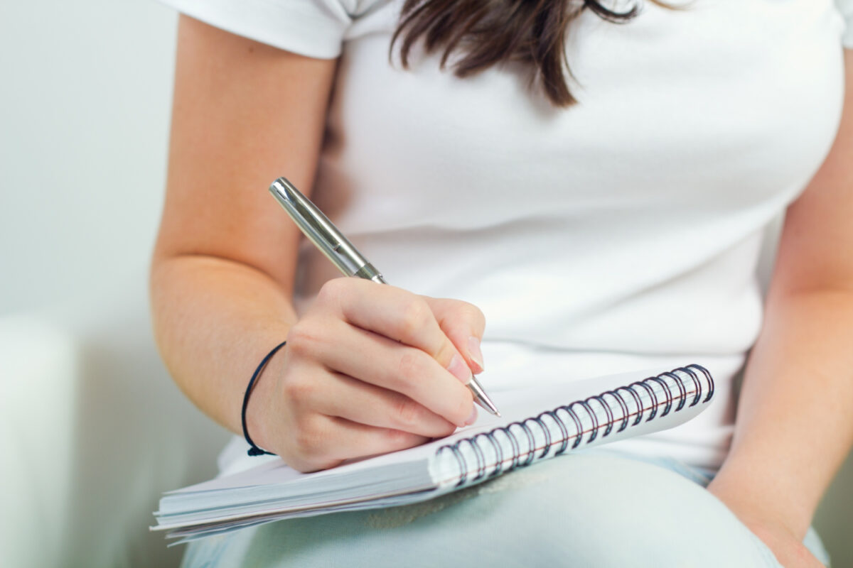 Mulher de blusa branca escrevendo em caderno espiral apoiado no colo