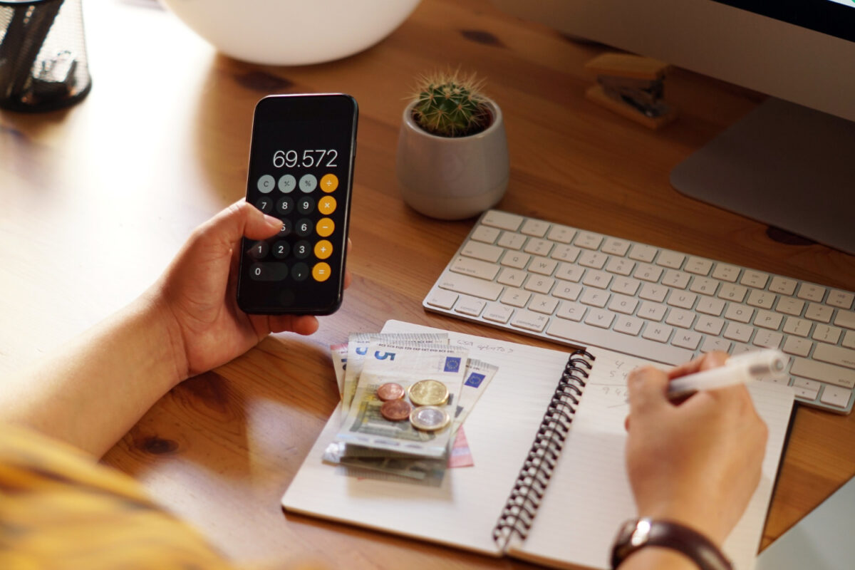 Mãos usando calculadora no celular ao lado de cadernos com anotações financeiras e cédulas sobre mesa de madeira
