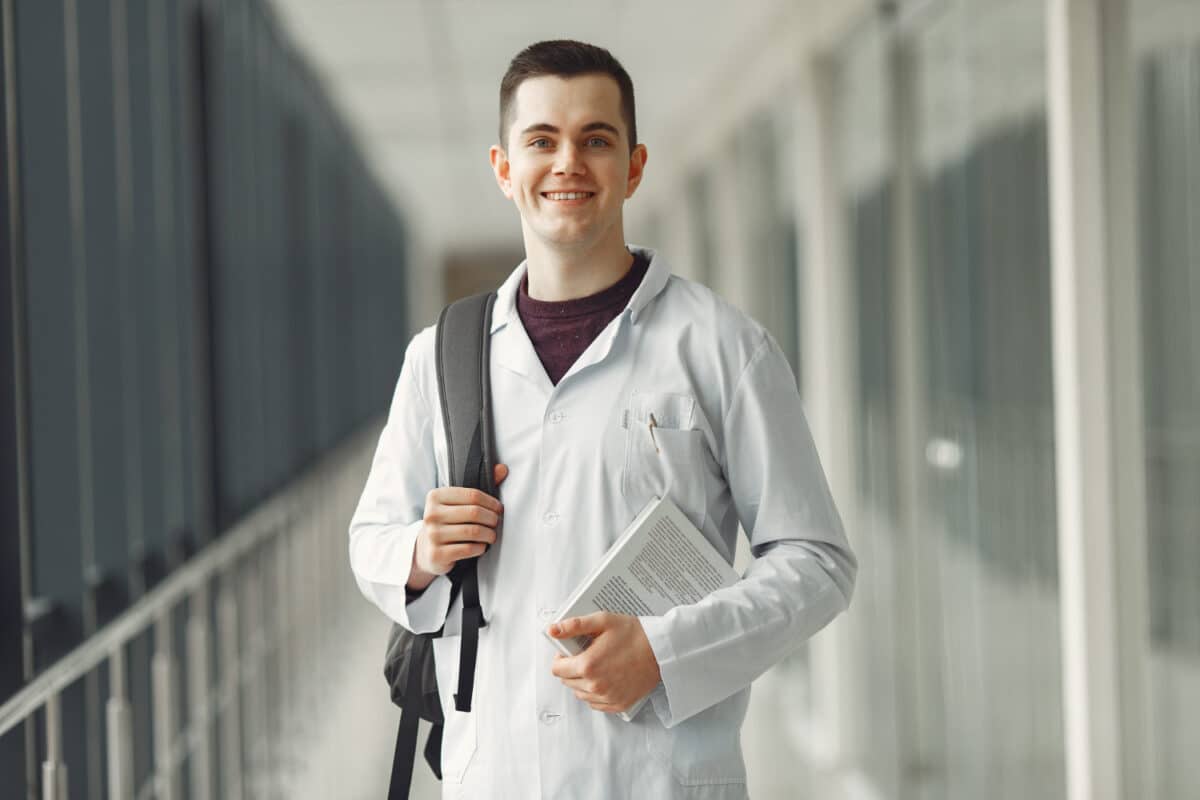 Estudante de medicina com jaleco branco e mochila sorri em corredor de instituição de ensino