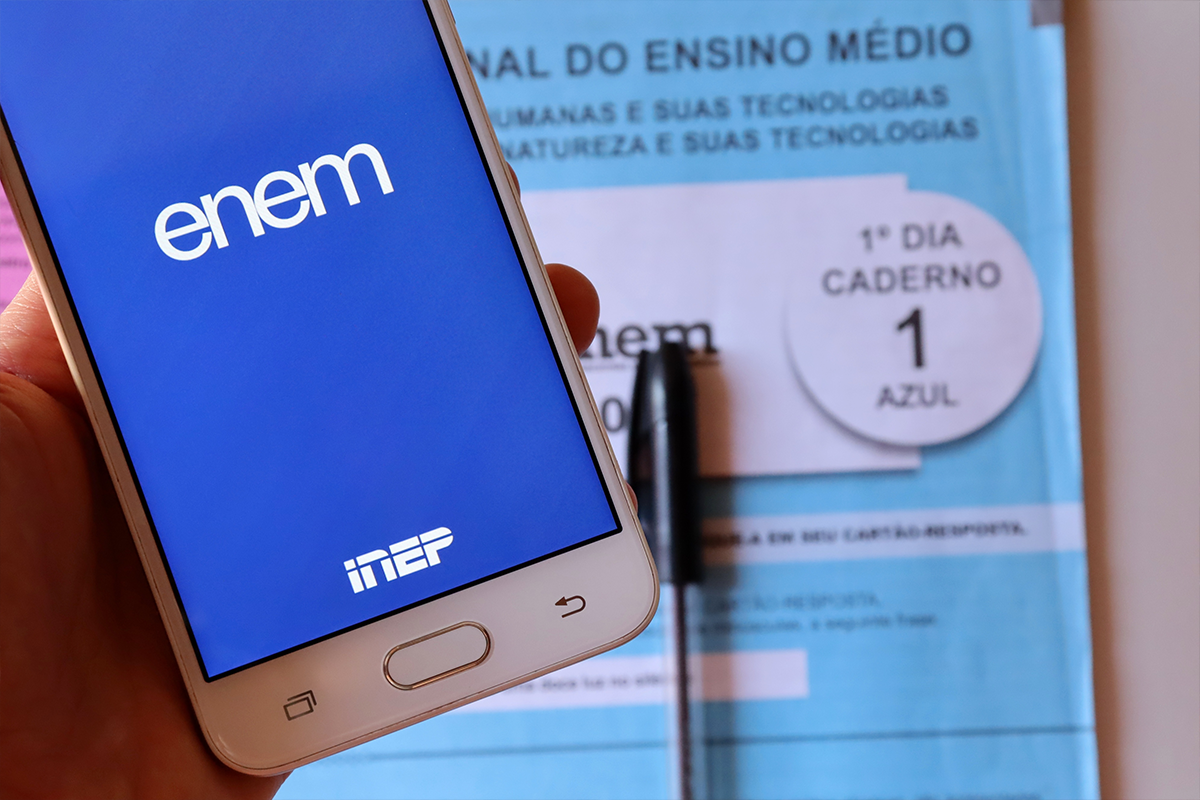 Mão segurando celular com app ENEM/INEP sobre caderno de prova azul do 1º dia de Ciências Humanas e Natureza.