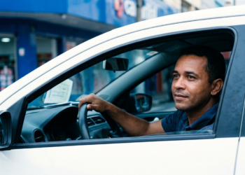 Motorista de aplicativo segura o volante dentro de carro branco estacionado em rua comercial