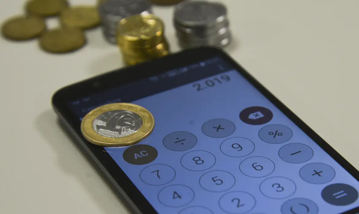 Smartphone com aplicativo de calculadora aberto na tela ao lado de pilhas de moedas brasileiras