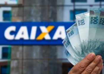 Mão segura cédulas de R$ 100 em frente à fachada de uma agência da Caixa Econômica Federal