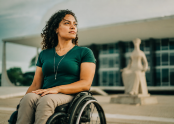 Mulher de cabelos cacheados em cadeira de rodas diante da estátua A Justiça, no STF, Brasília.