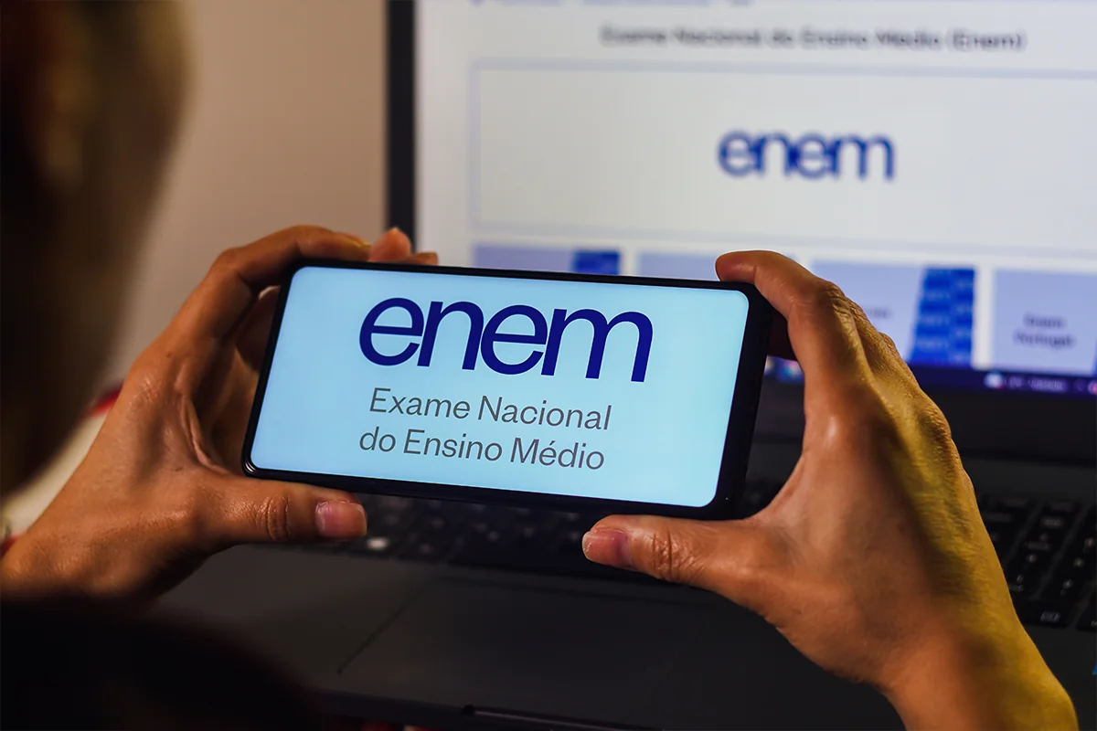 Pessoa acessa informações do Enem pelo celular, com tela de computador ao fundo, em contexto sobre resultado e recursos da isenção do Enem 2026.