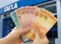 Mãos seguram em leque cédulas de dez, vinte, cinquenta e cem reais em frente a uma agência da Caixa.