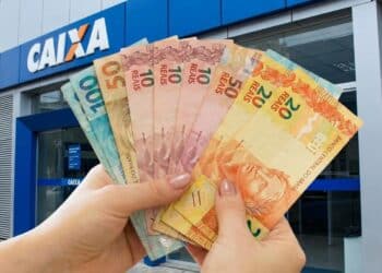 Mãos seguram em leque cédulas de dez, vinte, cinquenta e cem reais em frente a uma agência da Caixa.
