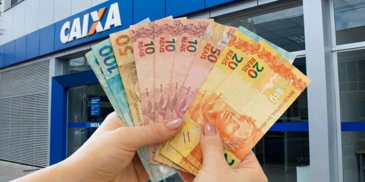 Mãos seguram em leque cédulas de dez, vinte, cinquenta e cem reais em frente a uma agência da Caixa.