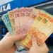 Mãos seguram em leque cédulas de dez, vinte, cinquenta e cem reais em frente a uma agência da Caixa.