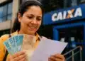 Mulher sorrindo, segurando notas de R$ 100 e R$ 50 em frente à agência da CAIXA, representando o saque de R$ 6.220 disponível até 1º de junho.