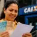 Mulher sorrindo, segurando notas de R$ 100 e R$ 50 em frente à agência da CAIXA, representando o saque de R$ 6.220 disponível até 1º de junho.