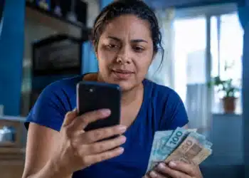 Mulher consulta no celular informações sobre o saque de R$ 6.220 da Caixa, representando a solicitação do valor, que pode ser feita até 30 de maio.
