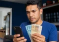 Trabalhador brasileiro consulta informações do FGTS no celular enquanto segura notas de reais em casa, representando o saque de R$ 6.220 do Fundo.