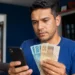 Trabalhador brasileiro consulta informações do FGTS no celular enquanto segura notas de reais em casa, representando o saque de R$ 6.220 do Fundo.