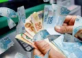 Notas de R$ 100 sobre imagem de dinheiro sendo sacado, representando o pagamento da segunda parcela do 13º salário do INSS.