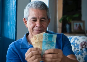 Aposentado brasileiro segurando notas de reais em casa, representando o pagamento da segunda parcela do 13º do INSS.