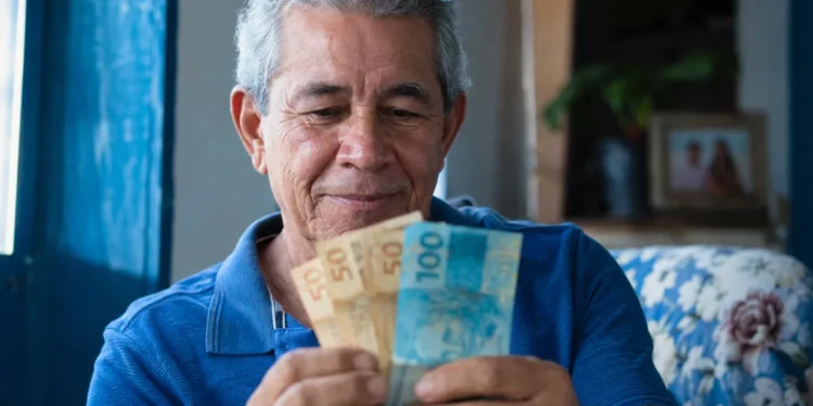 Aposentado brasileiro segurando notas de reais em casa, representando o pagamento da segunda parcela do 13º do INSS.