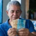 Aposentado brasileiro segurando notas de reais em casa, representando o pagamento da segunda parcela do 13º do INSS.
