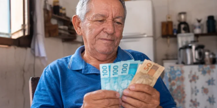 Aposentado brasileiro segurando notas de reais em casa, representando a liberação da segunda parcela do 13º salário do INSS