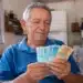 Aposentado brasileiro segurando notas de reais em casa, representando a liberação da segunda parcela do 13º salário do INSS